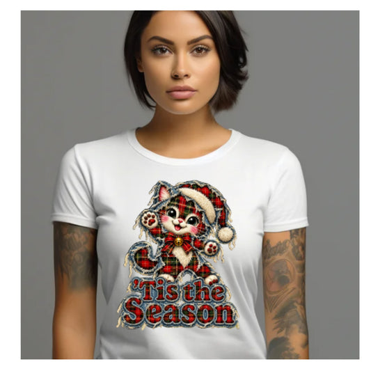A woman wearing a white short-sleeve crewneck t-shirt with a festive graphic of a plaid cat in a Santa hat and the text 'Tis the Season'.